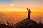 person standing on the top of the mountain with hand up, back view, over the city at sunset