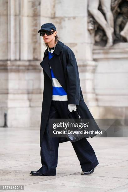 Simon Nygard wears a black cap, black sunglasses, silver earrings, a silver large chain necklace, a black high neck pullover, a white and royal blue...