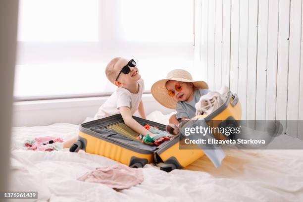 smiling happy children tourists a boy and a little girl in sunglasses and a hat are packing summer things in a suitcase going on vacation sitting on the bed at home. little brother and sister are having fun in anticipation of summer vacation and weekend - baby bag bildbanksfoton och bilder