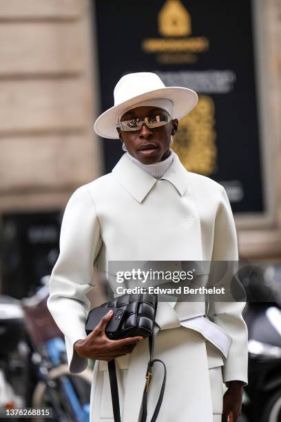 Guest wears a white felt / wool cap, black sunglasses, a white ribbed turtleneck pullover, a white belted long coat, a black shiny leather handbag...