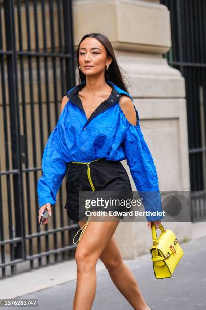 Guest wears gold earrings, a royal blue cut-out shoulder / oversized shirt, a black short skirt with yellow zipper, a yellow shiny leather handbag,...