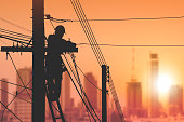 Silhouette electrician on ladder is installing cable lines on electric power pole with blurred cityscape view in sunrise sky background