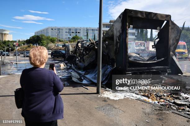 Pedestrian uses her smartphone to take footage of a damaged and burnt merchandise truck near an Aldi store in the Les Flamants neighbourhood, in the...