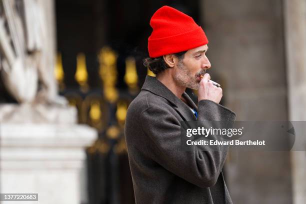Guest wears a red wool beanie, a blue and white large striped print pattern zipper neck pullover, a gray long coat, outside Nanushka, during Paris...