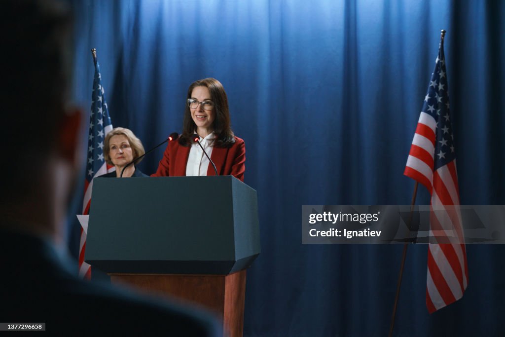 Dark haired female politician during the speech on a blue background with American flags