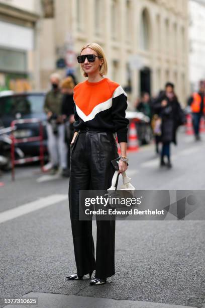 Lisa Aiken wears black sunglasses, a neon orange / white / black wavy large straps wool pullover, high waist black shiny leather large pants, a gold...