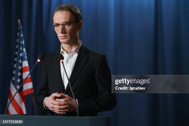 portrait photo of a serious young white politician looking away, standing against the blue background with american flag - microphone debate stock pictures, royalty-free photos & images