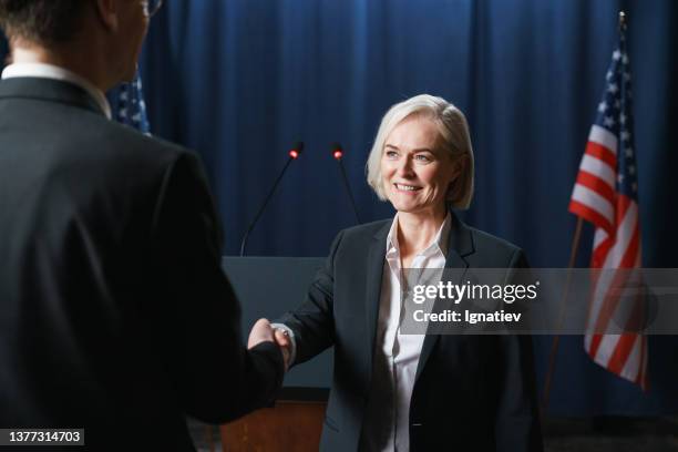 female american politician shakes a hand of her colleague, we see them on a blue background with american banner - us president office stock pictures, royalty-free photos & images