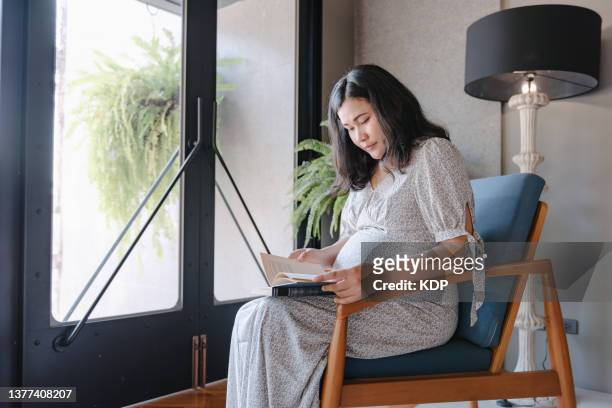 pregnant woman reading a book in living room. - maternity wear stock pictures, royalty-free photos & images
