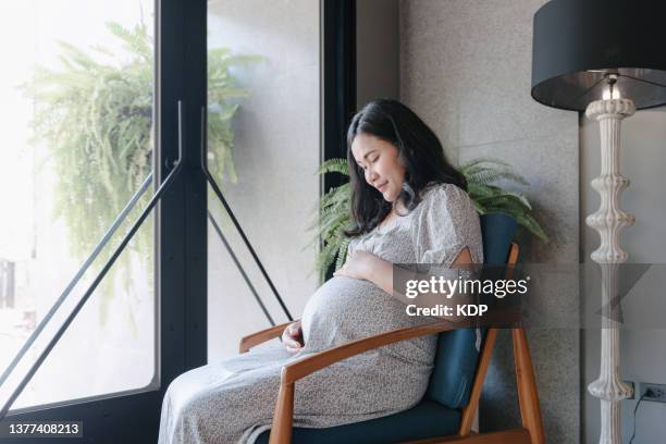 pregnant woman touching her belly while sitting for relaxing in living room. - maternity wear stock pictures, royalty-free photos & images