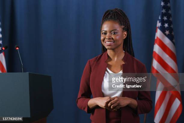 schöne afrikanische politikerin in einem roten anzug, die vor der rede mit einem lächeln vor der kamera posiert und vor dem blauen hintergrund mit amerikanischen flaggen steht - mayor stock-fotos und bilder