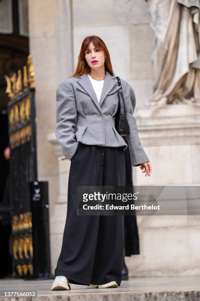 Guest wears gold necklace, a white ribbed t-shirt, a gray oversized jacket, a black shiny leather shoulder bag, black wide legs pants, beige sneakers...