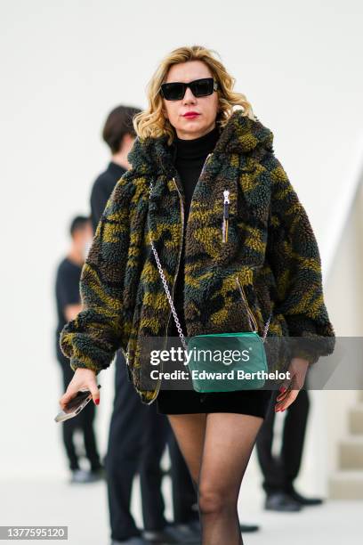 Guest wears black sunglasses from Prada, a black turtleneck pullover, a brown / green / khaki military print pattern oversized hoodie jacket, a green...