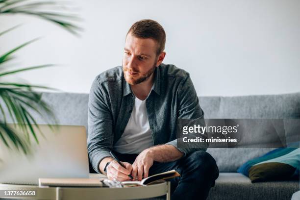 work at home: man sitting on sofa writing notes - project traveller stock pictures, royalty-free photos & images