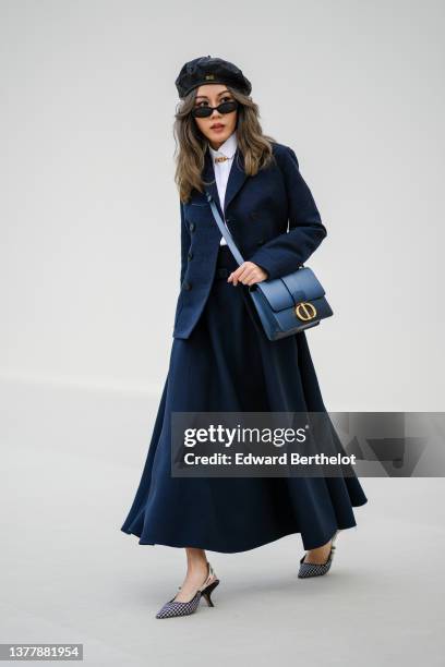 Guest wears a black shiny leather beret from Dior, black sunglasses, a white shirt, a gold large chain necklace from Dior, a navy blue jacket from...