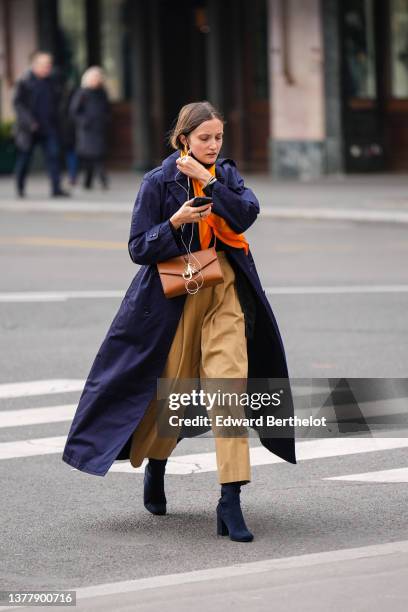 Guest wears gold earrings, an orange scarf, a black wool pullover, a navy blue oversized coat, gold rings, a brown shiny leather JW Anderson...