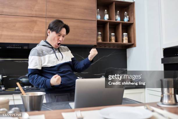 un uomo dell'asia orientale in un maglione ha applaudito mentre guardava il suo laptop nella cucina di famiglia. - 僅一男人 foto e immagini stock