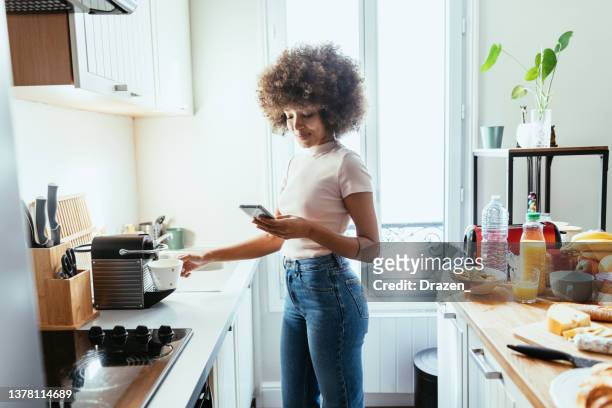 mixed race woman using smart phone in kitchen and preparing coffee from coffee machine - coffee chat stockfoto's en -beelden