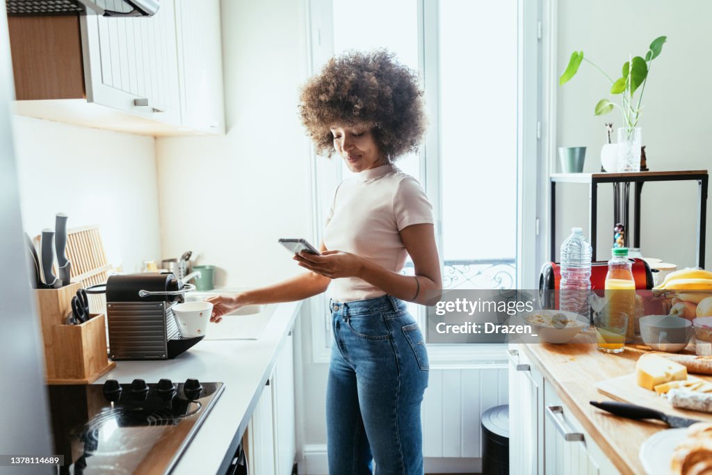 Mixed race woman using smart phone in kitchen and preparing coffee from coffee machine