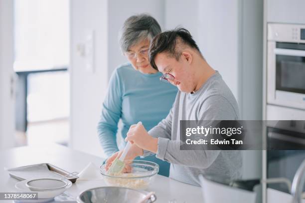 asiatisch-chinesischer down-syndrom-enkel lernt von seiner großmutter, die kuchen in der küche zubereitet - 2 people back asian stock-fotos und bilder