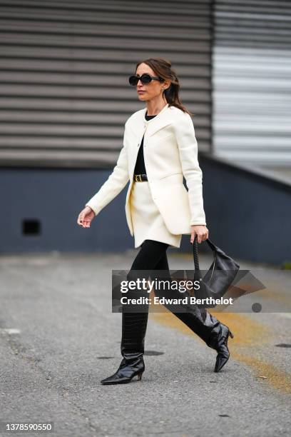 Chloe Harrouche wears black sunglasses, gold and silver earrings, a black t-shirt, a white latte jacket, a matching short skirt, a black shiny...