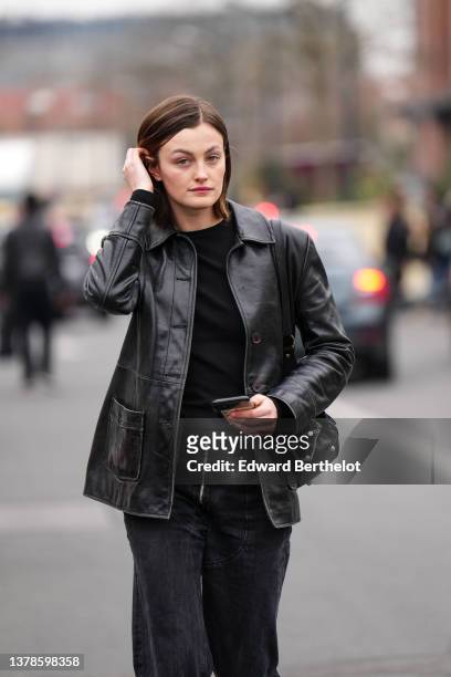 Model wears a black turtleneck pullover, a black shiny leather jacket, black large denim jeans pants, a black shiny leather shoulder bag, outside...