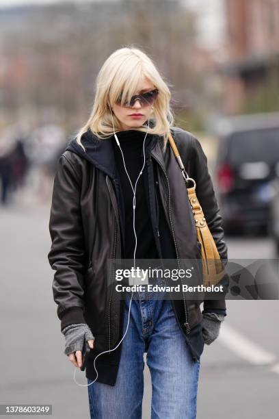 Model wears black sunglasses, gold earrings, a black high neck pullover, a black shiny leather zipper jacket, gray wool mittens, a black belt, blue...