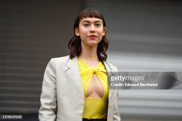 Guest wears a yellow knot neck / cut-out chest body from Coperni, a pale gray shiny leather with white cross print pattern long coat, a blue pearls...