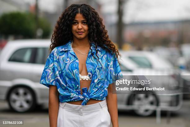 Guest wears gold and silver chain necklaces, a blue / purple / green / white print pattern short sleeves / knot / open shirt from Coperni, a white...