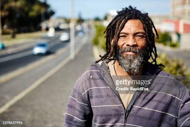homeless man with beard and dreadlocks outdoors in city in sunny weather - homeless person stockfoto's en -beelden