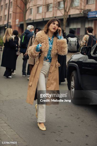 Jeanne Damas talks on the phone and wears a tan leather jacket with a shearling collar, blue print shirt, white pants, and light yellow ballet flats...