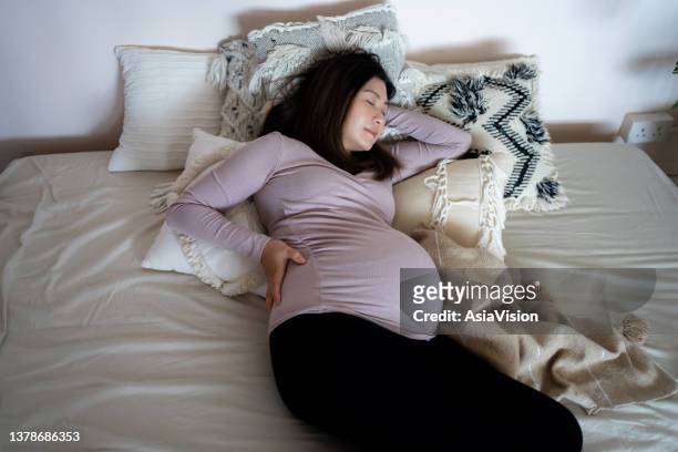 high angle shot of asian pregnant woman holding her belly, sleeping on bed. comfortable sleeping positions during pregnancy. insomnia, sleeping disorder and physical discomfort during pregnancy - back pain bed stock pictures, royalty-free photos & images