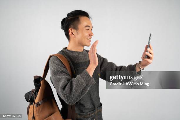 a young asian male, carrying a bag against a white background, smiles as he talks to a friend on his smartphone video - asiatischer mann freigestellt stock-fotos und bilder