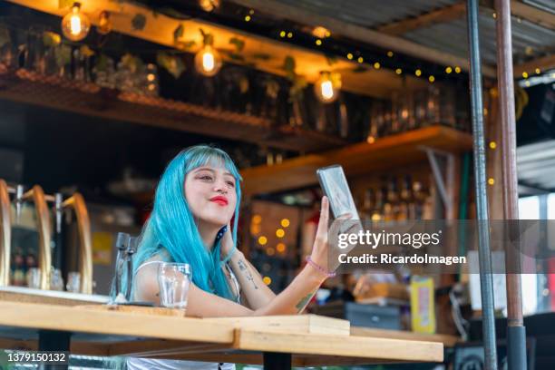 young latin woman with blue hair is sitting at a restaurant table taking a selfie with her cell phone - blue hair stock pictures, royalty-free photos & images