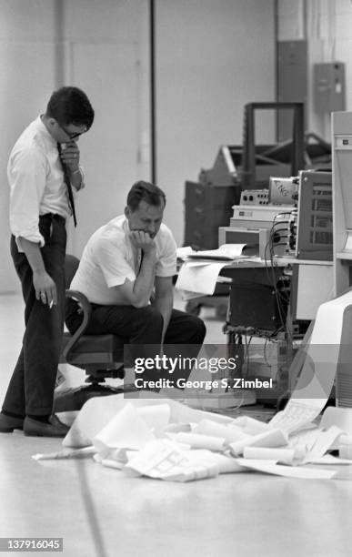 Two engineers look a ream of paper, piled on the floor from a printer, 1965.