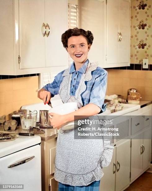 1950s Smiling Brunette Woman Housewife Wearing Apron In Kitchen Stirring A Bowl Of Batter Looking At Camera.