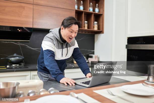 un uomo dell'asia orientale tiene un laptop in mano nella cucina di famiglia e il suo volto mostra un'espressione sorpresa - 僅一男人 foto e immagini stock