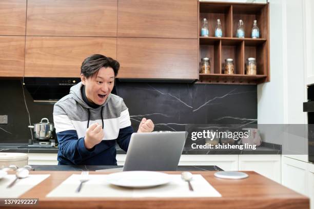 un uomo dell'asia orientale tiene un laptop in mano nella cucina di famiglia e il suo volto mostra un'espressione sorpresa - 僅一男人 foto e immagini stock