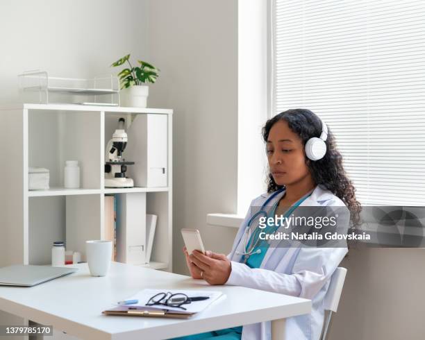 african-american doctor surfing the net on smartphone while listening to music in headphones - doctor listener imagens e fotografias de stock
