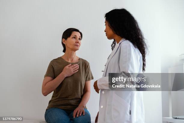 mature female patient explaining chest ache to african-american doctor in white coat - hand on female chest stock pictures, royalty-free photos & images