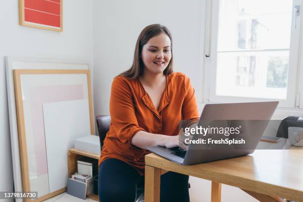 smiling plus size woman sitting at the table in her home office and typing something on her laptop - fat people stock pictures, royalty-free photos & images