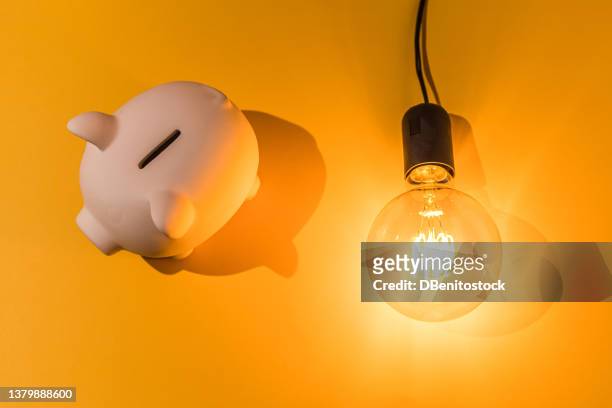 piggy bank next to lit light bulb on pink background. concept of electricity price, crisis, money, saving and power energy. - spaargeld stockfoto's en -beelden