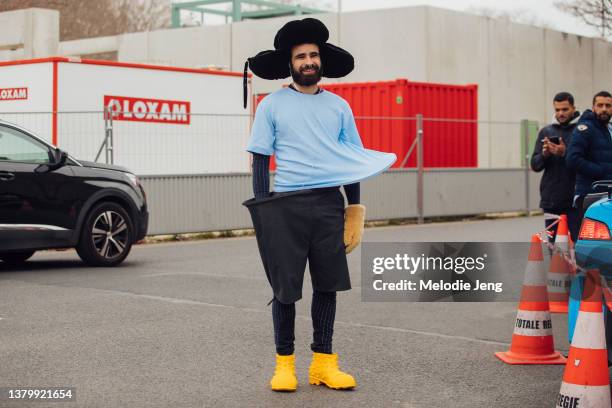 Ryan Benacer wears an oversized black hat with ears, Loewe asymmetrical oversized angular baby blue top, Loewe open shorts, black jeans, yellow...