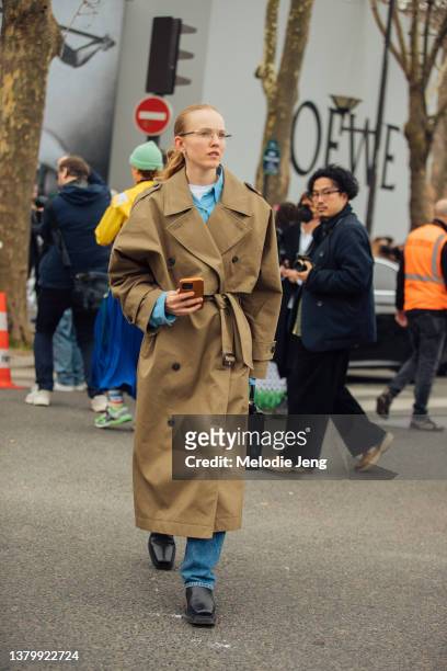 Stylist Alexandra Carl wears a belted tan trench coat, blue top, blue jeans, and black square-toe boots at the Loewe show at Tennis Club de Paris...
