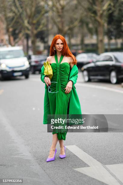 Guest wears green sunglasses, a yellow matte Flamenco clutch in nappa calfskin handbag from Loewe, a green long coat, a green V-neck / shoulder-off /...