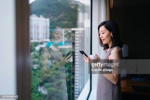 confident young asian businesswoman reading emails on smartphone and having coffee while standing in front of office window. female leadership. business lifestyle with technology - coffe house live stock pictures, royalty-free photos & images