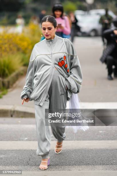 Guest wears gold pendant earrings, a pale gray zipper oversized with orange print pattern sweater from Loewe, matching pale gray sport pants from...