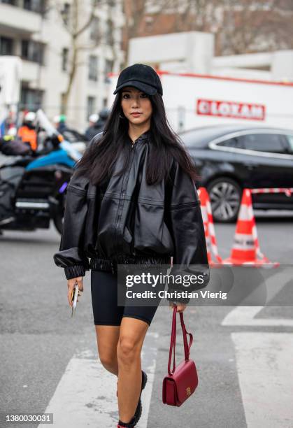 Yoyo Cao seen wearing black oversized leather jacket, cycle pants, red Loewe bag, laced boots, black cap outside Loewe during Paris Fashion Week -...