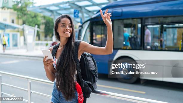 smiling asian female tourist with a mobile phone trying to call or searching the net, while waiting for a taxi - bus station stock pictures, royalty-free photos & images