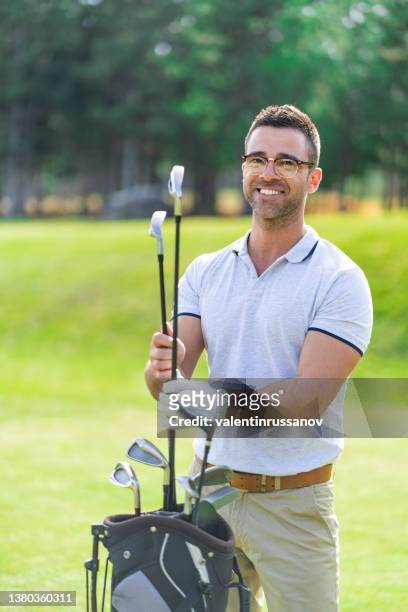 smiling golf player with eyeglasses, holding golf clubs, standing next to his bag on a golf course. - golf accessories stock pictures, royalty-free photos & images